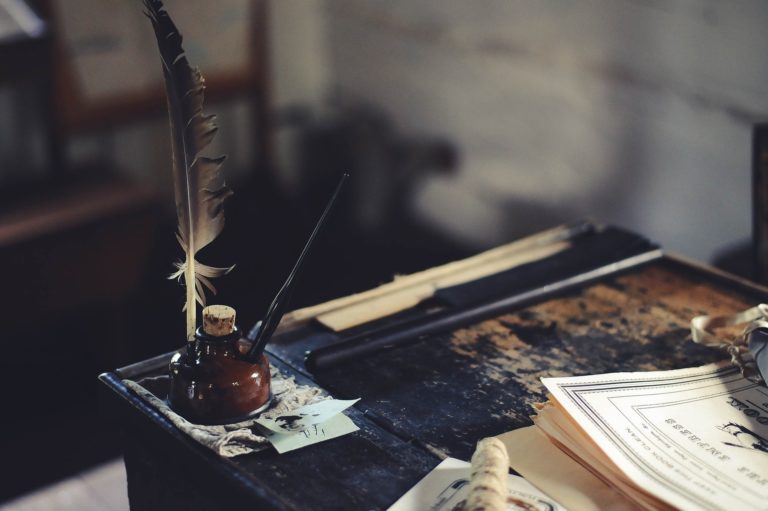 zdjęcie przedstawia kałamarz z atramentem oraz umieszczonym w nim ptasim piórem. Kałamarz jest umieszczony na biurku, obok widoczne są dokumenty.