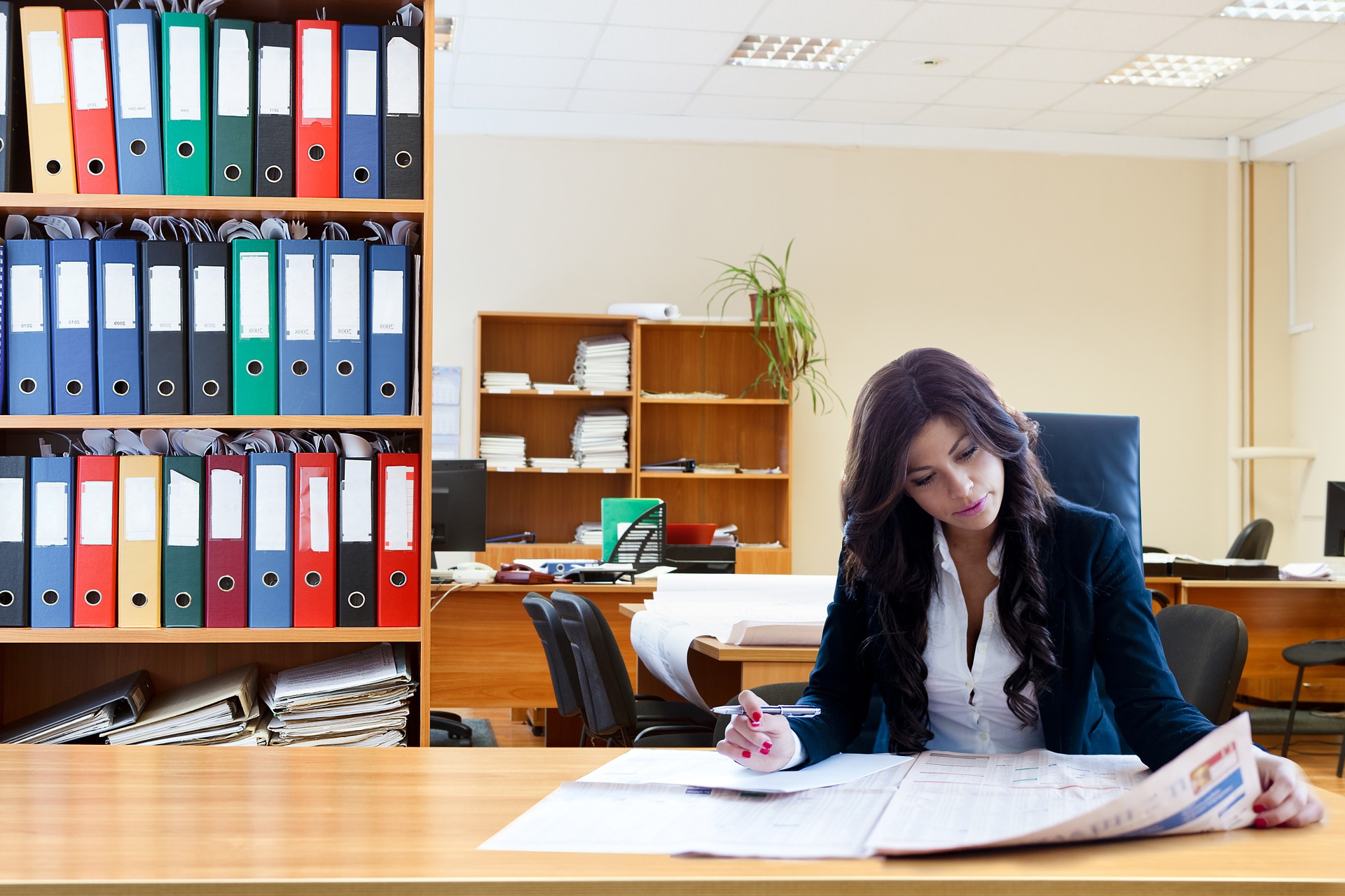 Zdjęcie przedstawia kobietę znajdującą się w pomieszczeniu biurowym; na drugim planie znajduje się przestrzeń biurowa w postaci regałów, biurek.